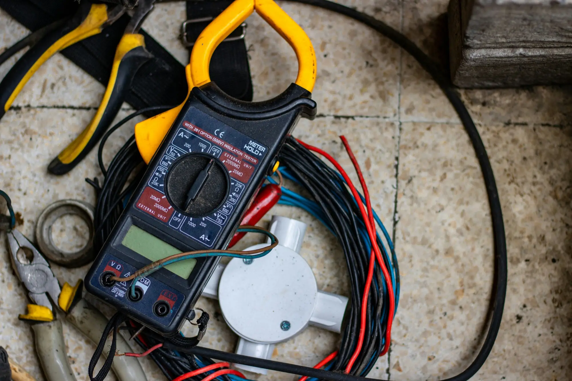 A yellow and black clamp meter with an insulation test range lying on a tiled floor among coils of wire, pliers and a junction box.