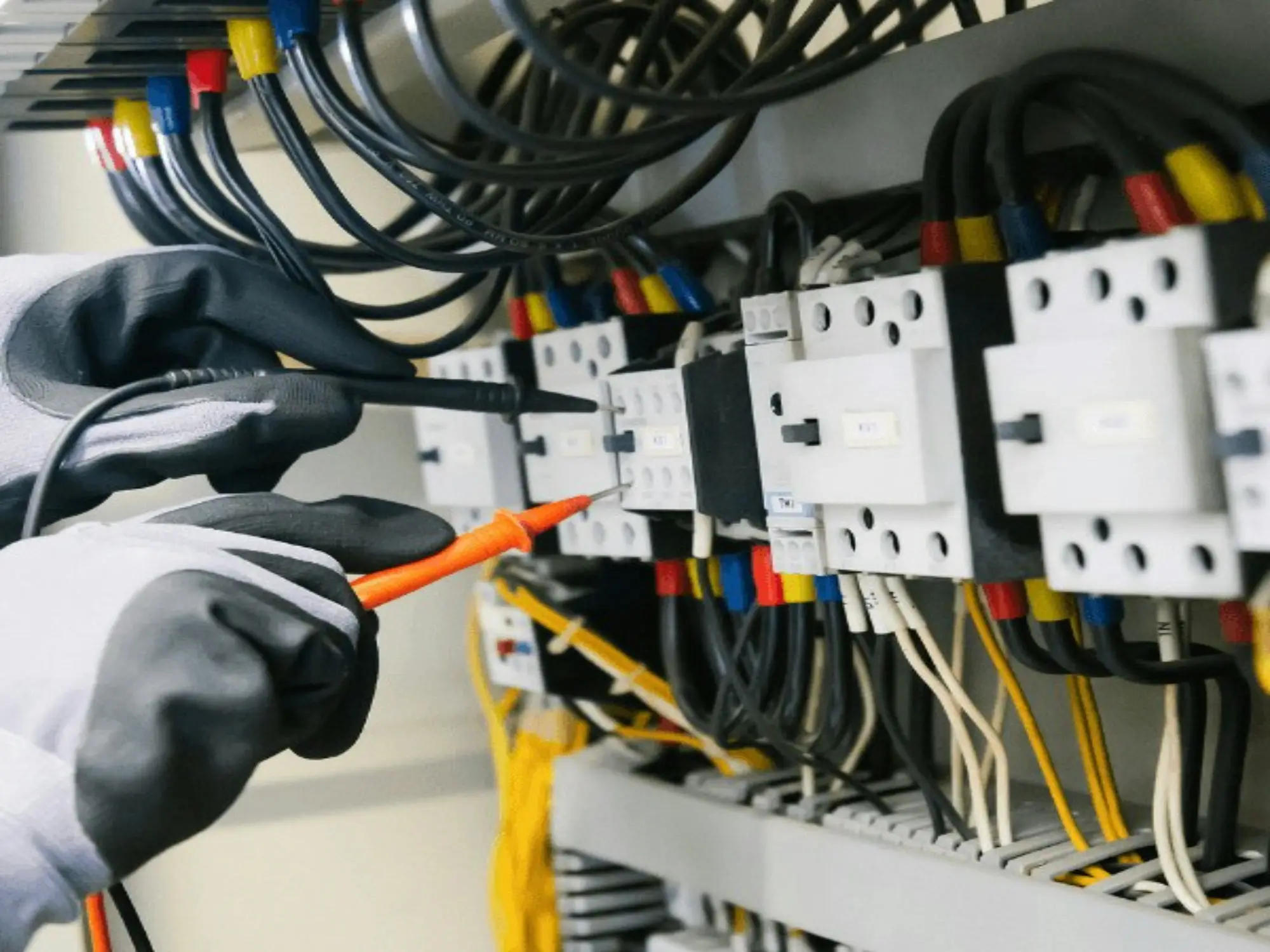 Gloved hands holding the two probes of a multimeter against the terminals of contactors in an electrical panel, with colour-coded wires above.