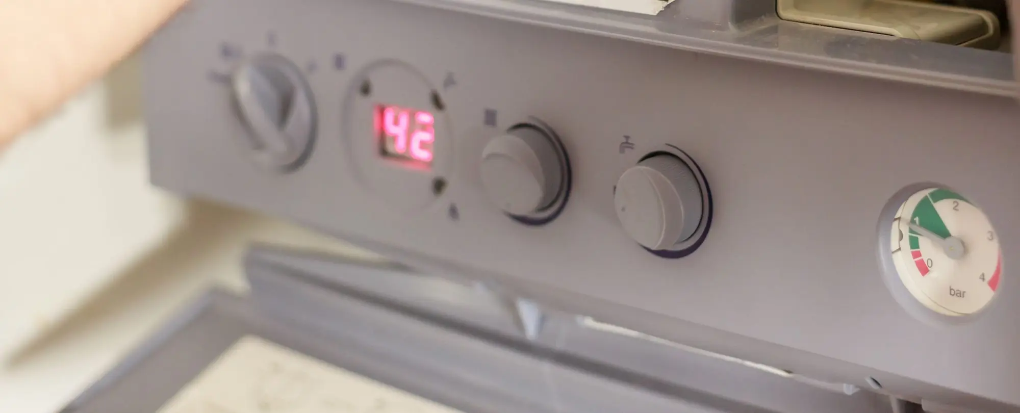 The control panel of a wall-mounted gas boiler: a temperature knob, a red digital display reading 42, two dials and a pressure gauge in bar, with an engineer's arm at the edge.